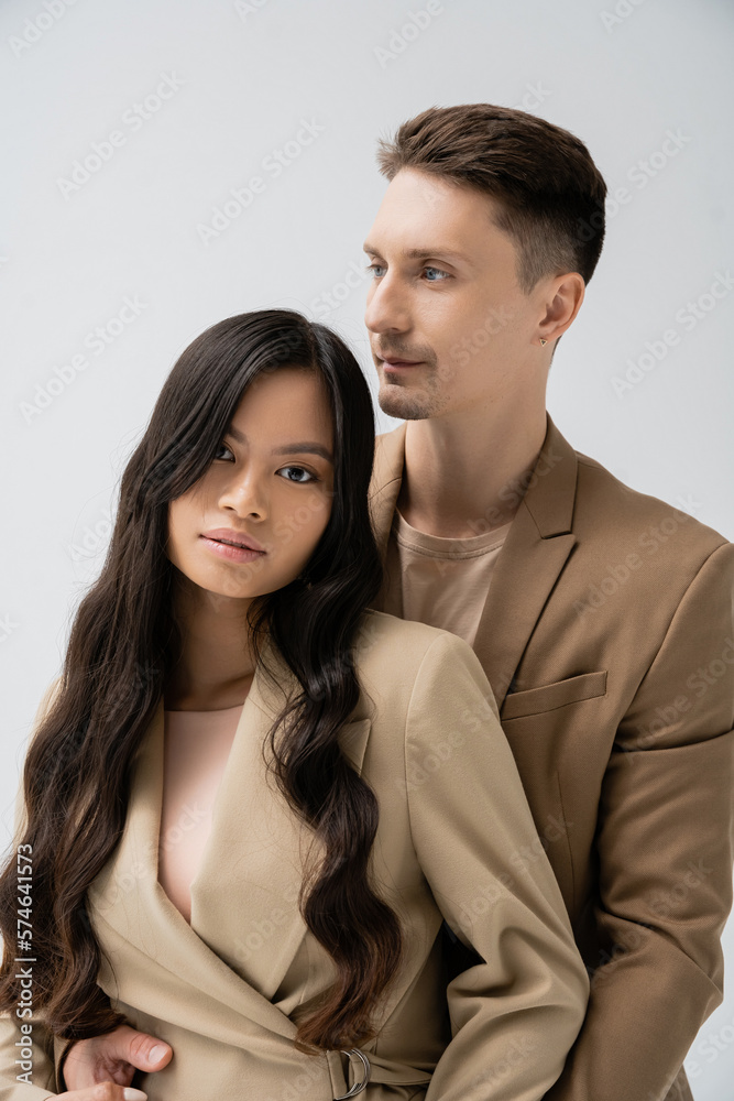 trendy man embracing pretty asian woman with long brunette hair isolated on grey. trendy man embracing pretty asian woman with long brunette hair isolated on grey.