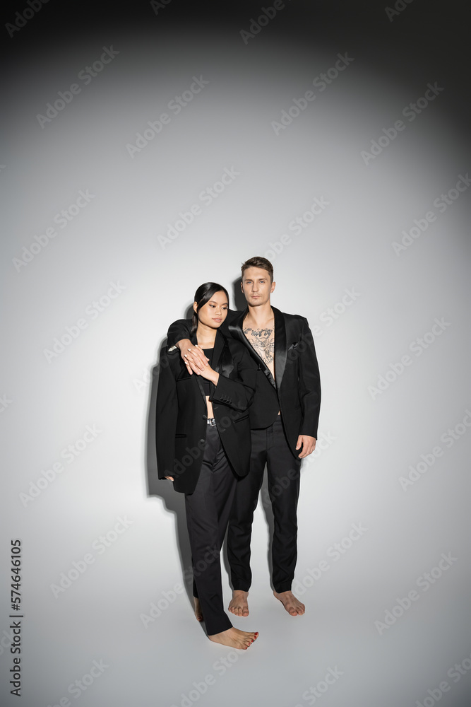 full length of barefoot multiethnic couple in black stylish pantsuits standing on grey background. full length of barefoot multiethnic couple in black stylish pantsuits standing on grey background.