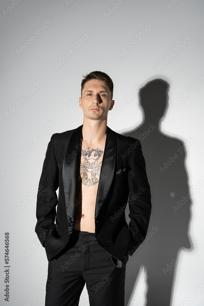 front view of shirtless tattooed man in black elegant pantsuit posing with hands in pockets on grey background with shadow. front view of shirtless tattooed man in black elegant pantsuit posing with hands in pockets on grey background with shadow.