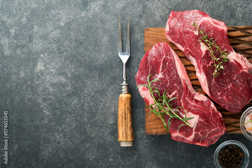 Raw beef steak. Marbled raw fresh Ribeye steak with rosemary, salt and pepper on wooden cutting board on dark concrete background. Raw beef steak and spices for cooking. Mock up.