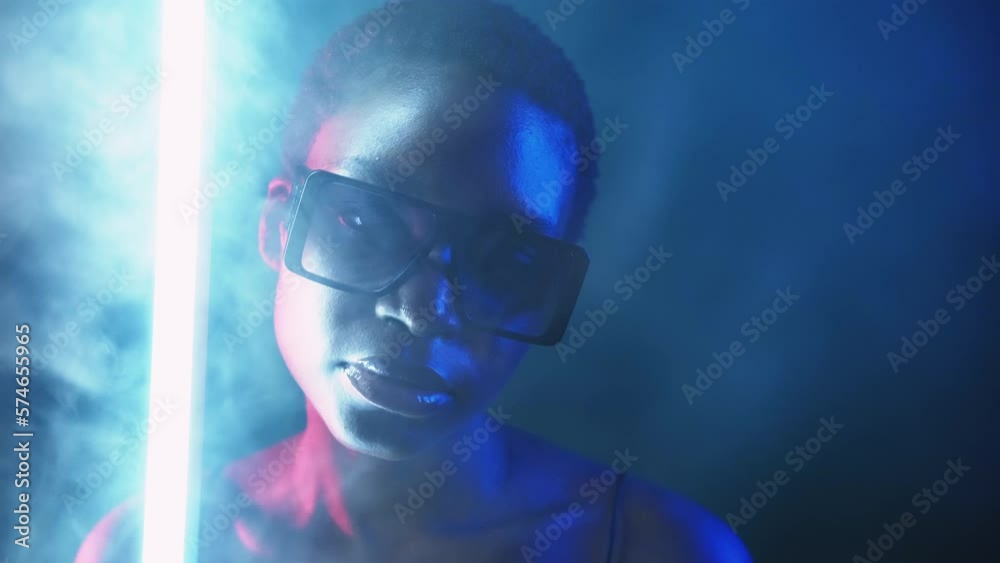 Eyewear fashion. Neon light face. Cyberpunk beauty. Blue red color mysterious girl posing in stylish sunglasses in smoke cloud at dark night with white LED lamp illumination.