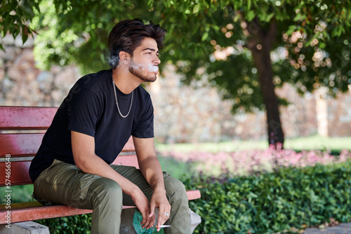 Attractive indian man smoker exhales cigarette smoke portrait in black t shirt and silver neck chain sitting on bench in public park, hindu male smoking close up portrait. Handsome indian man portrait photo