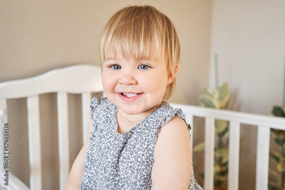  Happy baby girl one and half standing in the crib. Smiling 18 months child playing in sunny bedroom. Baby cot in the parent's bedroom