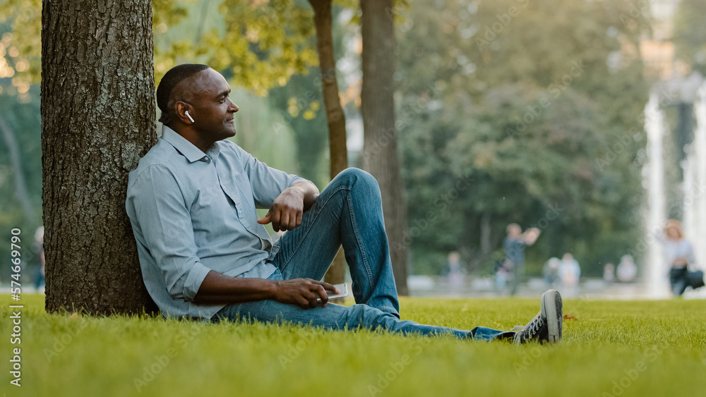 Happy calm relaxed elderly middle-aged male listening music with earphones mature senior adult African American ethnic man in wireless headphones sitting on grass in city park enjoy audio song sound
