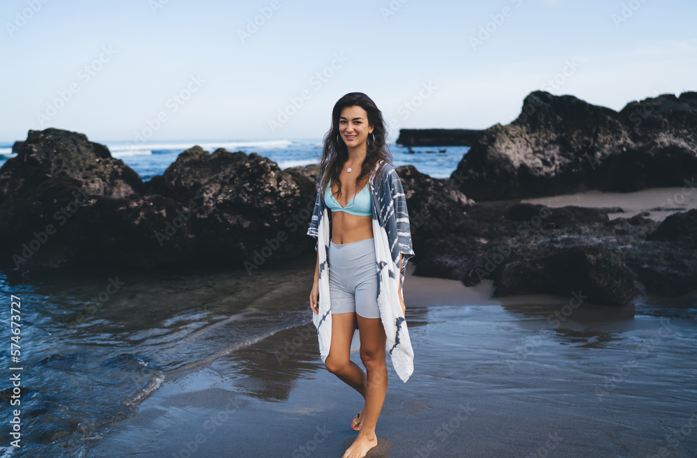 Positive ethnic barefooted lady standing on wet sandy seashore and smiling