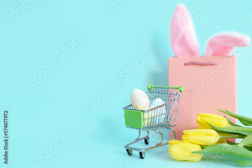 Easter eggs in a grocery basket on a blue background with yellow tulips and rabbit ears in a paper bag. easter, shopping. High quality photo