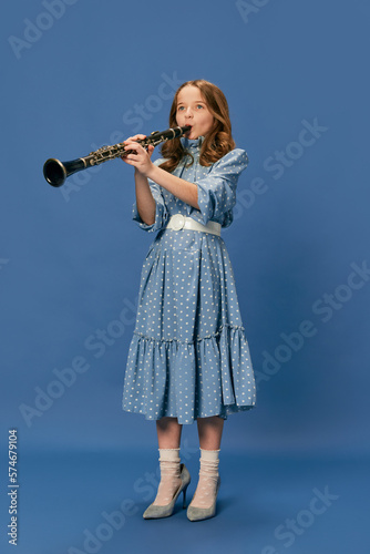 Monochrome portrait of charming nice little girl in dress and big shoes playing on clarinet over blue background. Music, fashion, hobby concept