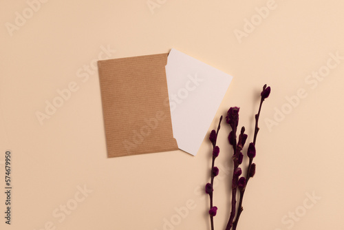 Mockup of an open invitation card made from eco-friendly kraft paper and decorated with a composition of dried flowering branches painted in burgundy color, arranged vertically on a beige background