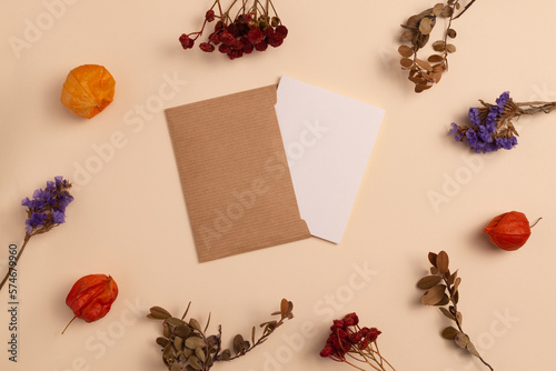 Mockup of an open greeting card with an envelope, made of eco-friendly kraft paper and decorated with a composition of dry branches and flowers, arranged around the card on a beige background
