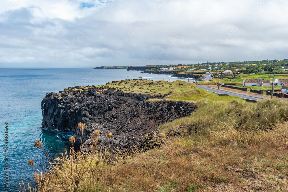 Volcanic rock cliff by the sea with dry herbs overlooking coastline and village, Fenais da Luz - São Miguel, Azores PORTUGAL