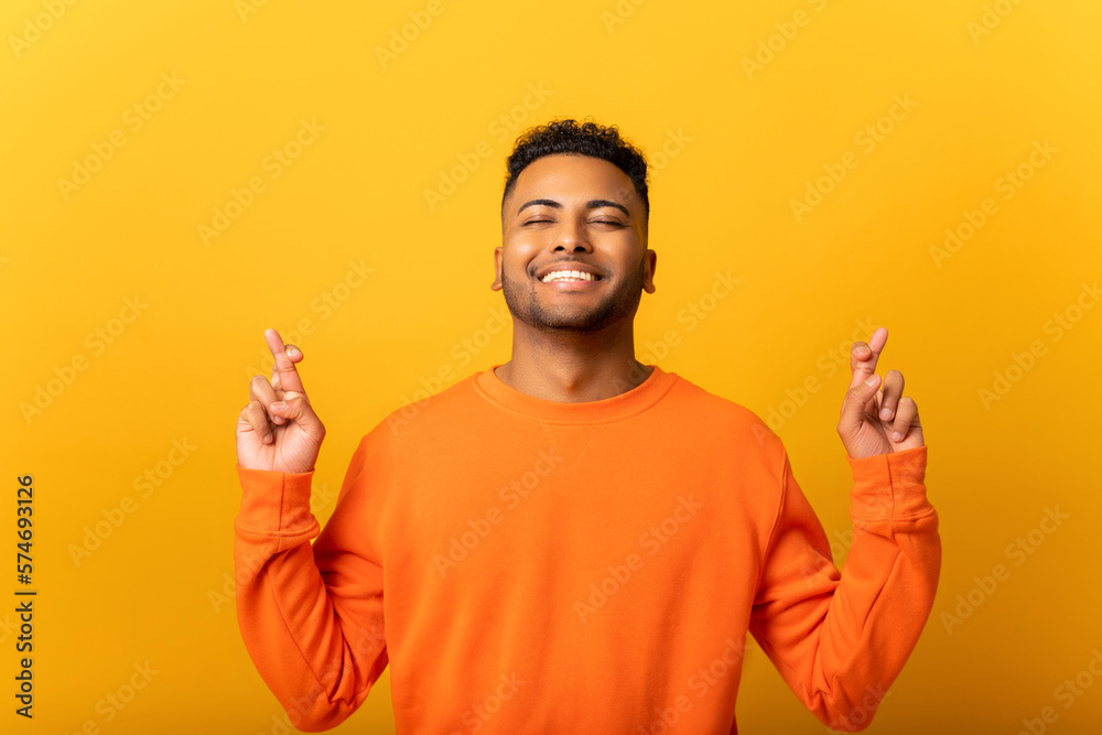 Wish you luck. Smiling young Indian man holding fingers crossed, looks up in hope, multiracial guy really wants to be lucky, isolated on yellow wall