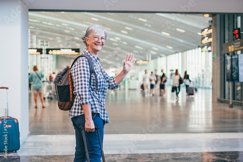 Happy attractive senior woman walking in airport area in direction to departure gate waving hand. Traveler vacation concept people with backpack and suitcases.