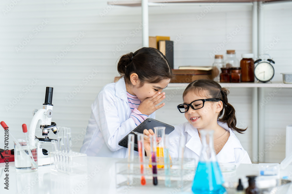 Two little Asian girls in lab coat whispering excitedly and smile together with many laboratory tools on shelves and table. Waiting for teacher checking result experiment in chemistry classroom. Two little Asian girls in lab coat whispering excitedly and smile together with many laboratory tools on shelves and table. Waiting for teacher checking result experiment in chemistry classroom.
