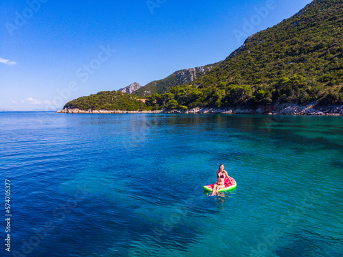 a girl in a bikini swims on a watermelon-shaped mattress with beautiful green mountains in the background  relaxing by the adriatic in the peljesac peninsula in croatia