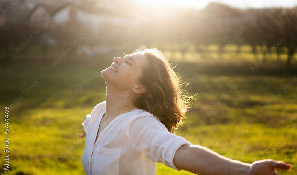 Young caucasian woman enjoying the sun and summer in a green field in the rays of the sun with her arms wide open - obrazy, fototapety, plakaty 