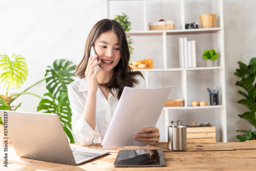 Beautiful Asian female accountant working with laptop at her desk analyzing business reports with folders, files and documents. Mobile smartphone messaging or text chatting. online work concept Beautiful Asian female accountant working with laptop at her desk analyzing business reports with folders, files and documents. Mobile smartphone messaging or text chatting. online work concept