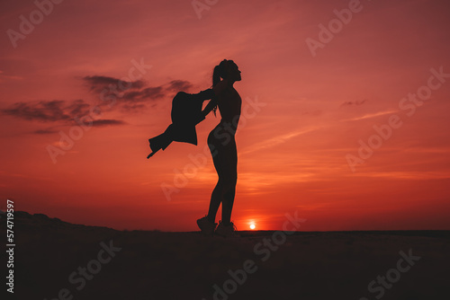 The girl is standing on top of the mountain and is holding the jacket she took off. A tourist on the background of a red beautiful sunset. Silhouette of a slender girl. © Alex