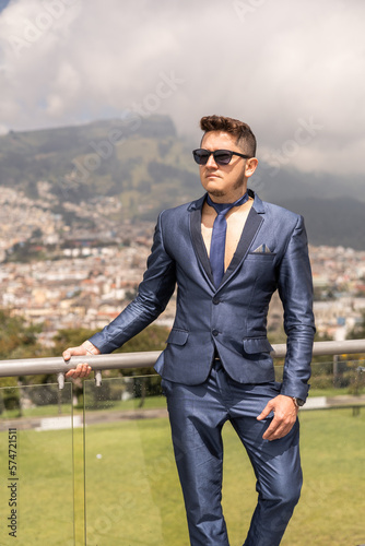 posing standing young man with short hair wears a blue formal suit, tie and sunglasses, lifestyle and business fashion, city landscape background photo