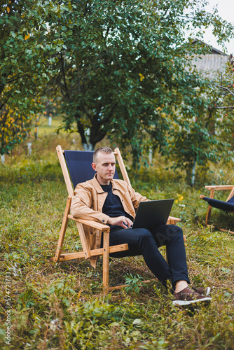A handsome man in a brown shirt sits on a wooden folding chair in the garden and works with a laptop. Wooden ecological furniture. Remote work © Дмитрий Ткачук