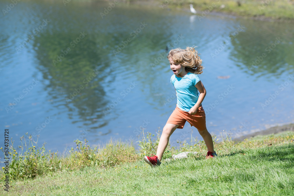 Kid boy running on green grass near lake in summer park. Kid boy running outdoor. Sport for children. Runner kids race in park. Kid boy running on green grass near lake in summer park. Kid boy running outdoor. Sport for children. Runner kids race in park.