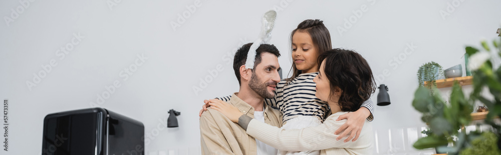 Fototapeta premium Smiling woman and daughter hugging man with Easter headband in kitchen, banner.