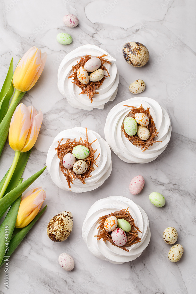 Easter treat - set of white meringues in shape of nest with multicolored candy chocolate eggs, tulips and quail eggs over marble background. Overhead view, flat lay. Holiday symbol