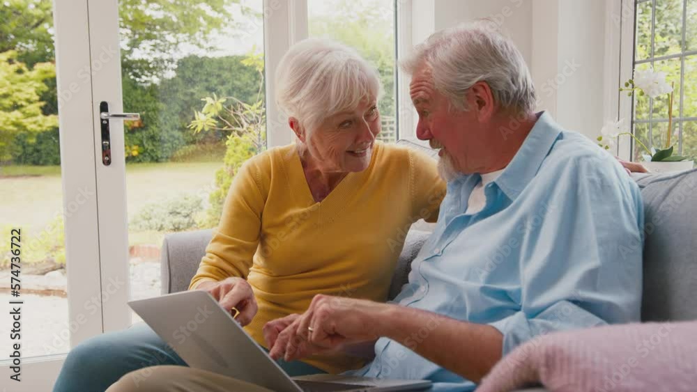 Excited retired senior couple with laptop sitting on sofa in lounge shopping online or booking holiday - shot in slow motion