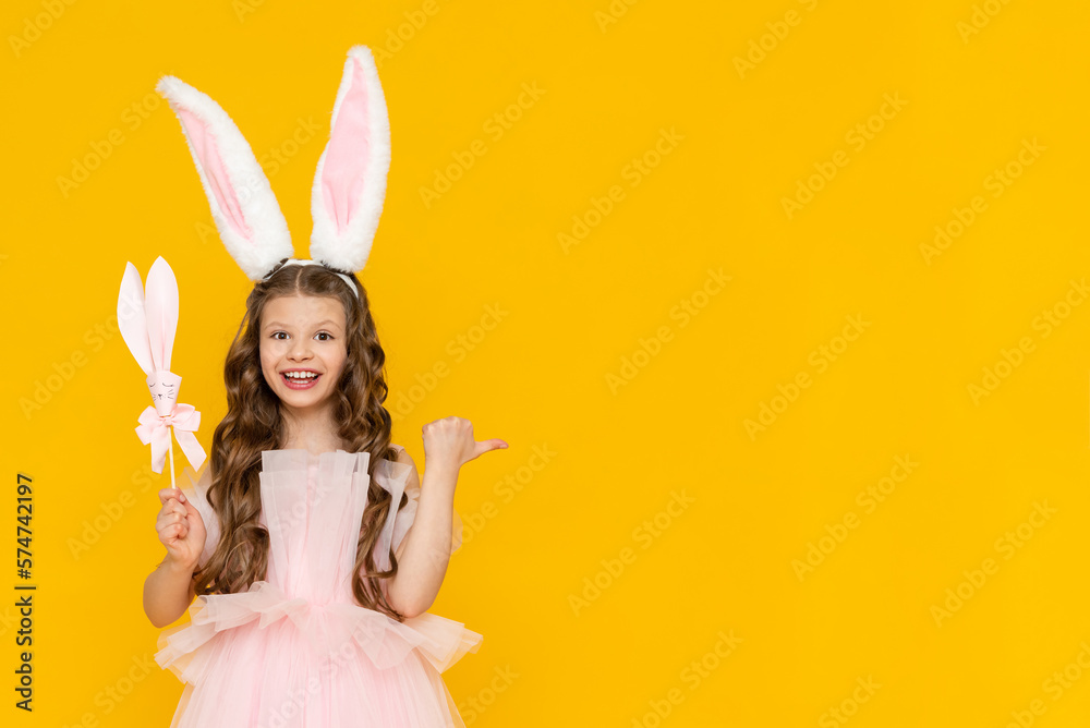 The little lady is celebrating Easter.  A beautiful little girl with curly hair is enjoying the spring holiday. A child with rabbit ears on his head, on a yellow isolated background. Copy space. 
