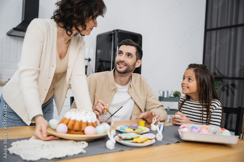 Smiling family coloring eggs near woman purring plate with Easter cake on table at home. Smiling family coloring eggs near woman purring plate with Easter cake on table at home.