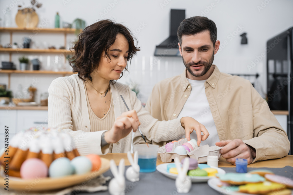 Woman holding paintbrush and taking Easter egg near husband in kitchen. Woman holding paintbrush and taking Easter egg near husband in kitchen.