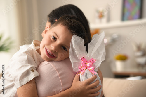 Joyful brazilian child looking at camera and hugging mother with easter egg in the hand at home photo