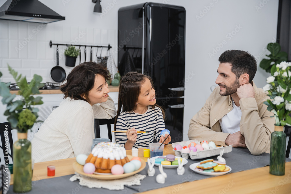 Smiling woman and daughter looking at man near Easter eggs and cake at home.