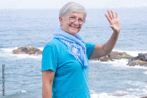 Portrait of smiling senior woman at sea looking at camera waving hand. Elderly retired female enjoying free time and vacation #574748552