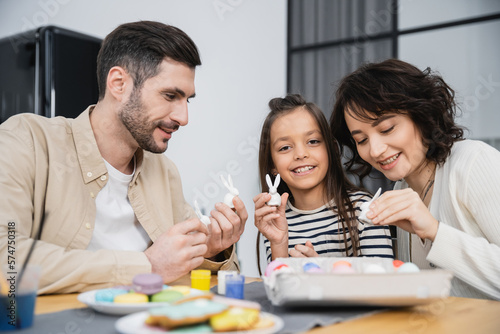 Smiling kid holding Easter rabbit figurine near parents and eggs at home.