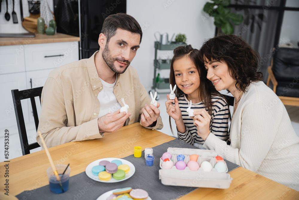 Parents and child holding Easter decor near eggs and macaroons in kitchen.