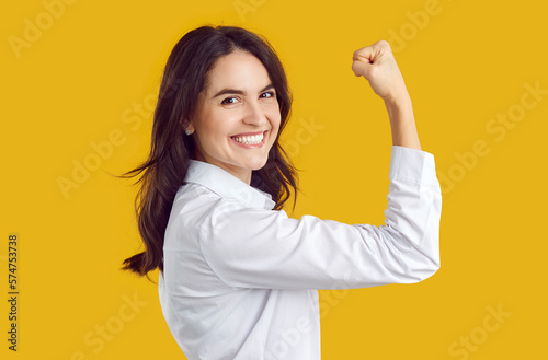 Joyful confident young woman show power gesture demonstrating her strength and leadership. Smiling brunette Caucasian woman in white shirt showing biceps on vivid orange background. Close up. photo