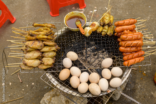 Metal grill with roasted chicken wings and feet placed near sausages and potatoes on sticks against eggs and bowl of sauce on brazier on Vietnam street