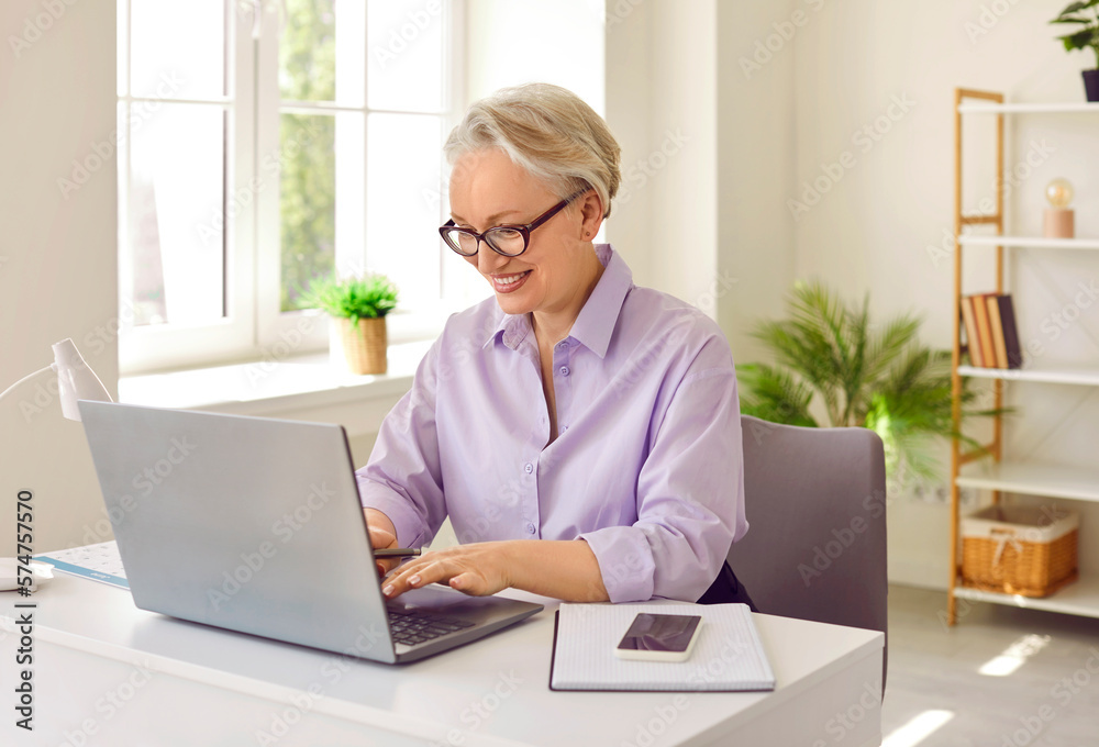 Positive mature elderly woman teacher learn schoolchildren and students via Internet in laptop answers questions in online chat and remotely helps to receive education sits in home office Positive mature elderly woman teacher learn schoolchildren and students via Internet in laptop answers questions in online chat and remotely helps to receive education sits in home office