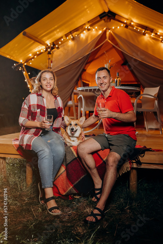 Happy lovely couple with Welsh Corgi Pembroke dog relaxing in glamping on evening and drinking wine near cozy bonfire. Luxury camping tent for outdoor recreation and recreation. Lifestyle concept © bondvit