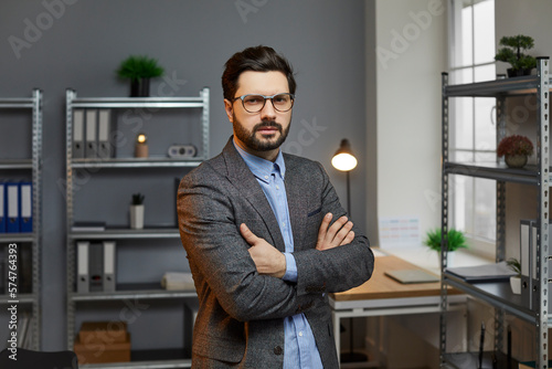 Portrait of serious confident businessman posing with arms crossed. Successful handsome bearded man wearing glasses and grey suit standing with arms folded looking at camera