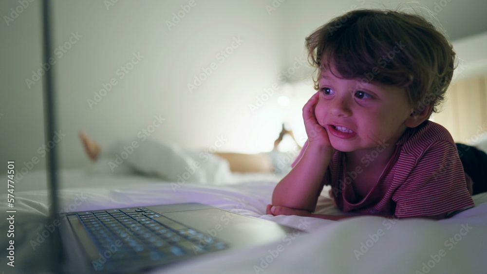 Young boy starring at media entertainment on laptop screen laying in bed at night. Hypnotized kid staring at blue glowing screen in the evening Young boy starring at media entertainment on laptop screen laying in bed at night. Hypnotized kid staring at blue glowing screen in the evening