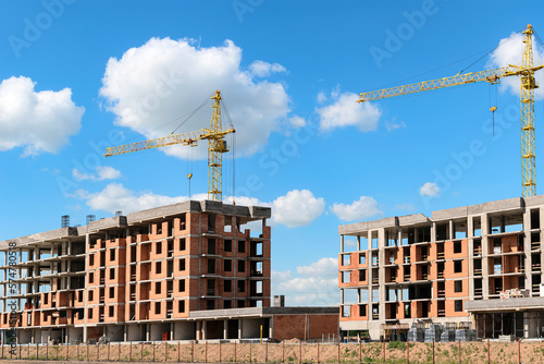 Construction site with multi-storey residential buildings under construction and cranes against a blue sky with clouds.