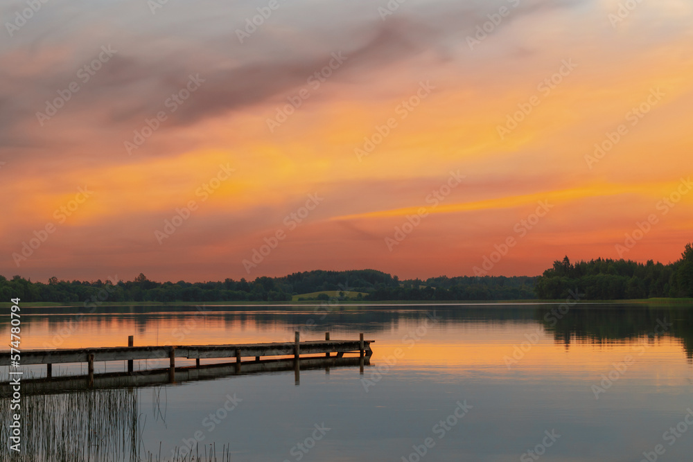 A view of a beautiful lake with a long wooden boardwalk. Lake landscape in summer evening sunset with orange sky