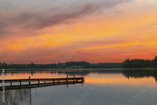 A view of a beautiful lake with a long wooden boardwalk. Lake landscape in summer evening sunset with orange sky