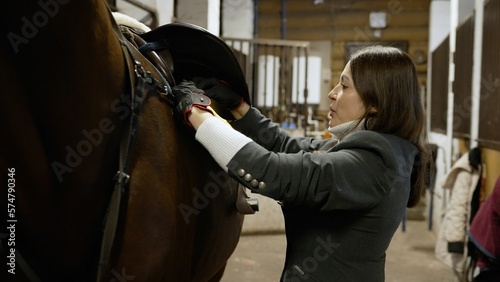 The girl attaches the saddle to the horse. A young woman saddled his horse for riding. Indoors in the stable.