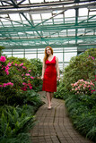 beautiful smiling romantic redhead girl in red dress walking in botanical garden with azaleas in a greenhouse