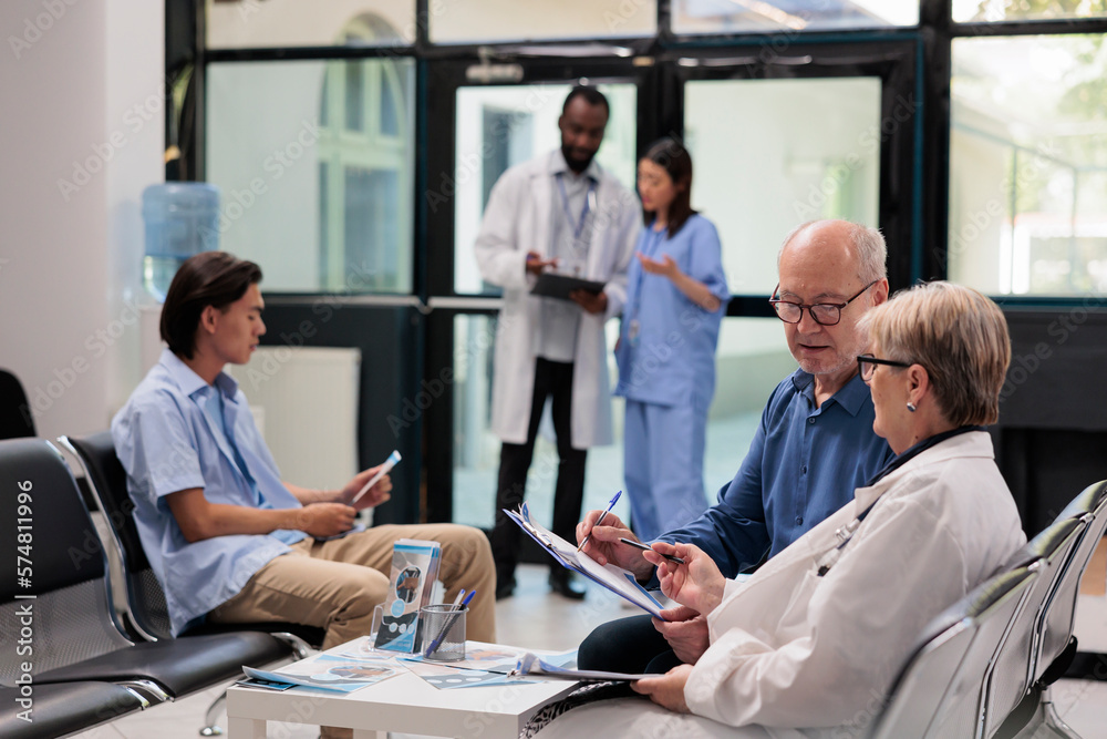 Obraz premium Senior specialist sitting on chair in hospital lobby explaining illness diagnosis to elderly patient discussing health care treatment. Diverse people waiting to start examination during checkup visit