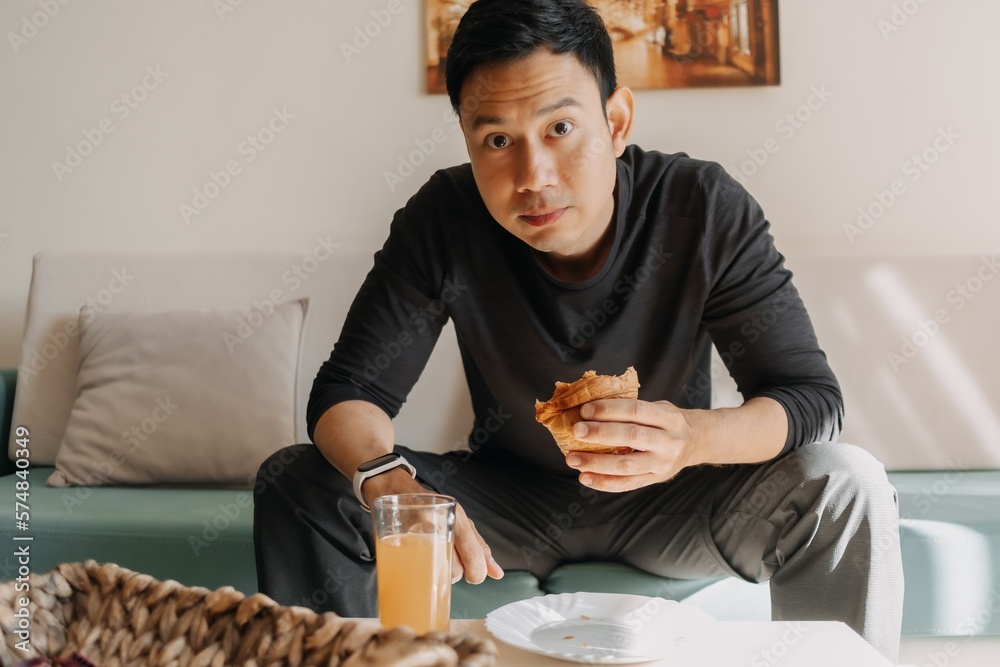 Asian man having bread and juice as breakfast on the sofa living room. Asian man having bread and juice as breakfast on the sofa living room.