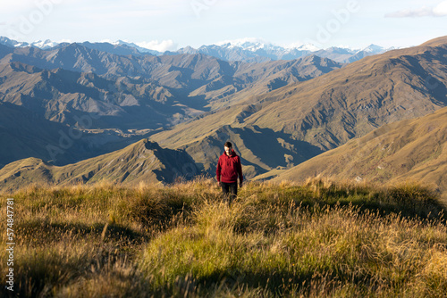 A young male hiker in a red sweater is walking on a tussock covered hill, with endless mountain peaks and snow mountains in the background at sunset. (ID: 574847781)