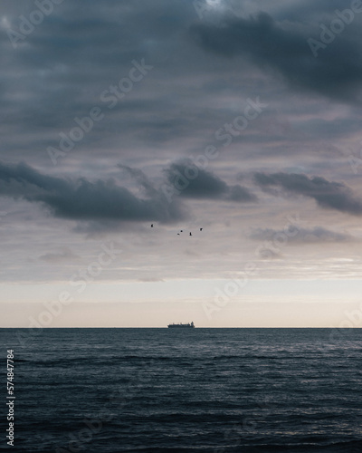 In the vast expanse of the ocean, a lone cargo ship cuts through the waves, its silhouette a stark contrast against the moody twilight sky. (ID: 574847784)